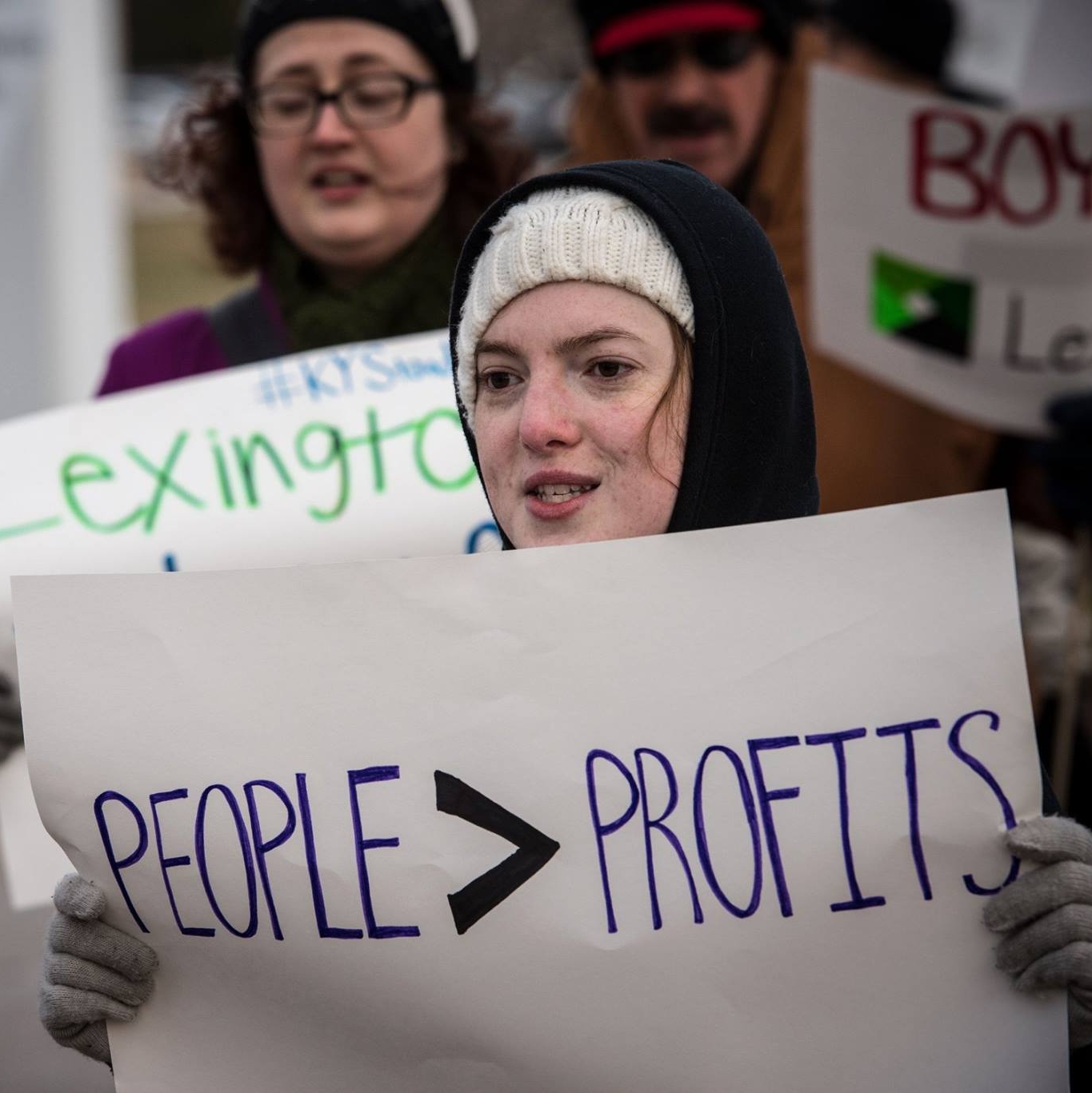 Robin Kunkel holding a sign that reads People > Profits, put differently people over profits. image credit Steve Pavey